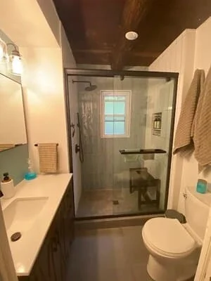 Bathroom with frameless glass shower, dark tile, white vanity, and updated lighting